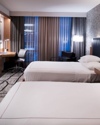 The image shows a modern hotel room with two neatly made beds, a wall-mounted TV, a desk with a chair, and stylish decor and lighting.