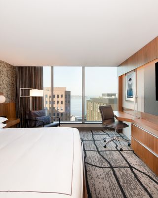 The image shows a modern hotel room with a large bed, a desk and chair, a flatscreen TV, and a view of city buildings through large windows.