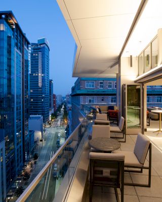 A modern balcony with seating overlooks a cityscape featuring tall buildings and a street below, illuminated at dusk.