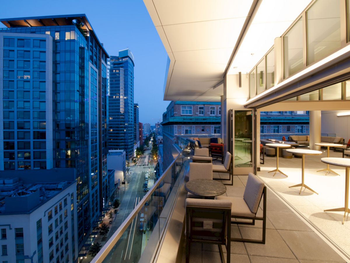 The image shows a modern cityscape with tall buildings and a balcony at dusk. The balcony has chairs, tables, and is well-lit.