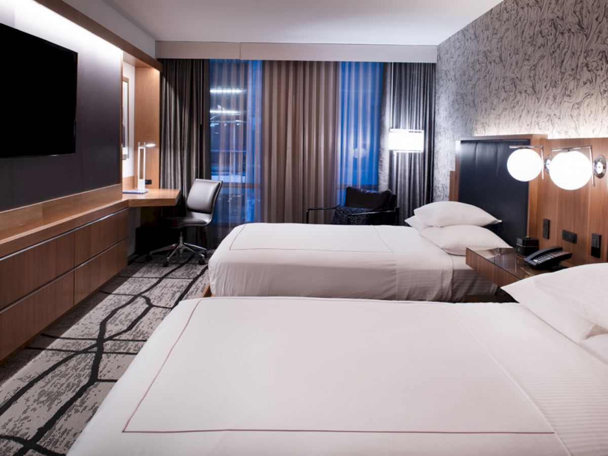 A modern hotel room with two beds, a flat-screen TV, a desk, and an illuminated window with curtains.