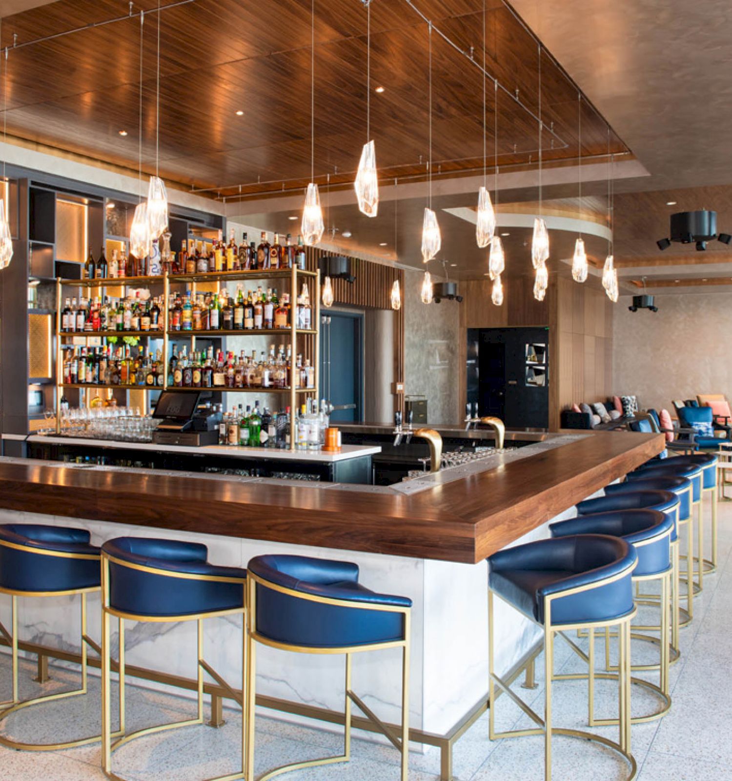This image shows a modern bar with a wooden counter, blue bar stools, hanging lights, and a well-stocked liquor shelf in a chic, spacious setting.