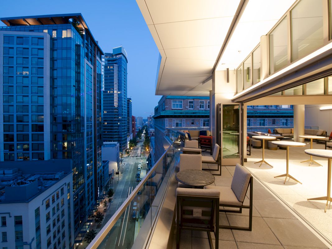 A modern balcony with seating and tables overlooks a city's skyscrapers and illuminated streets during the evening.