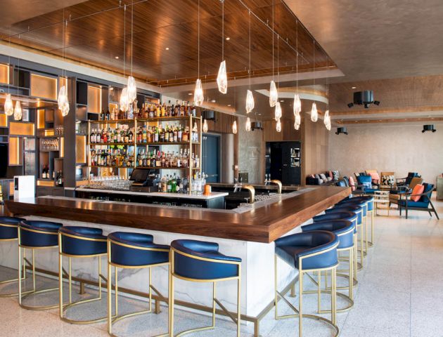 This image shows a modern bar with a wooden counter, blue bar stools, hanging lights, and bottles of liquor displayed behind the counter.