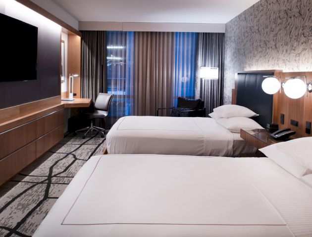 A modern hotel room with two beds, a flat-screen TV, a desk with a chair, and large windows with curtains.