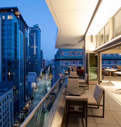 The image features a modern cityscape at dusk, viewed from a balcony with seating and tables. The buildings are lit up, and the street is visible below.