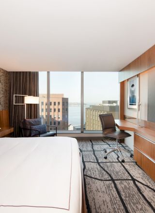 A modern hotel room with a king-sized bed, a large flat-screen TV, a desk, a chair, a patterned wall, and a view of nearby buildings through large windows.