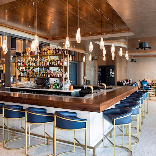 The image shows a modern bar with a wooden counter, blue stools, hanging light fixtures, and a well-stocked liquor shelf, located in a cozy setting.