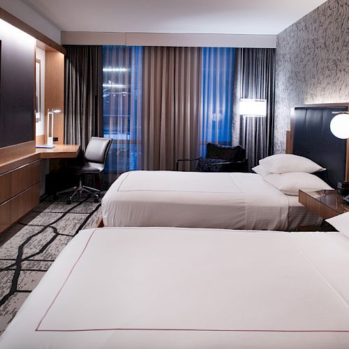 A modern hotel room with two beds, a wall-mounted TV, a desk with a chair, and contemporary decor including a patterned wall and floor.