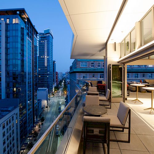 A modern cityscape with tall buildings and a wide balcony with tables and chairs overlooking a street at dusk.