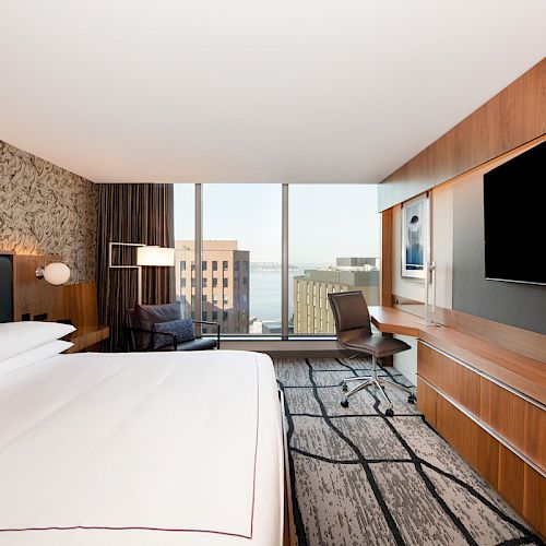 A modern hotel room with a large bed, a desk, a chair, a flat-screen TV, and a view of buildings and water through the window ends the sentence.
