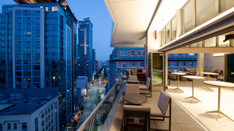A modern cityscape view from a spacious balcony at dusk, featuring chairs, tables, and buildings with glass facades lining the street.