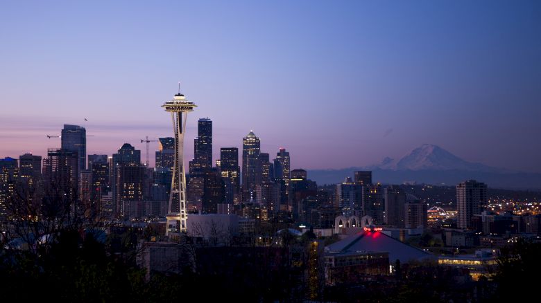 This image shows a city skyline at dusk with a prominent tower and mountain in the background, lights illuminating the buildings.