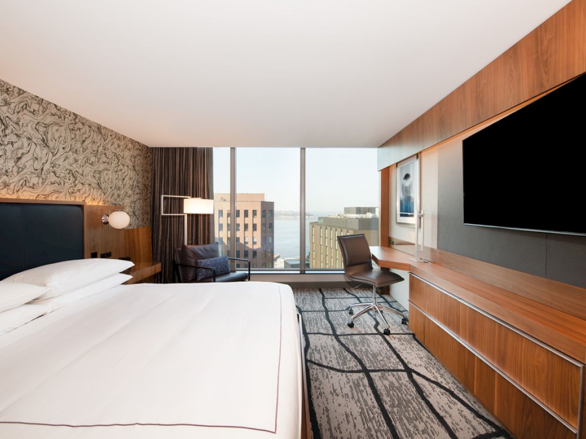 The image shows a modern hotel room with a large bed, a desk, chair, and a large TV. The room has a view of city buildings and a water body.