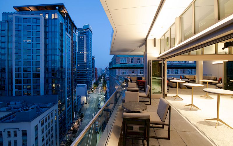A modern cityscape at dusk with tall buildings and a well-lit, stylish balcony featuring seating and tables overlooking the urban scene.