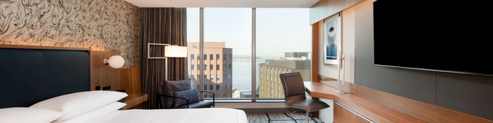 A modern hotel room with a large bed, a desk, a chair, a television, and a large window revealing a cityscape view.
