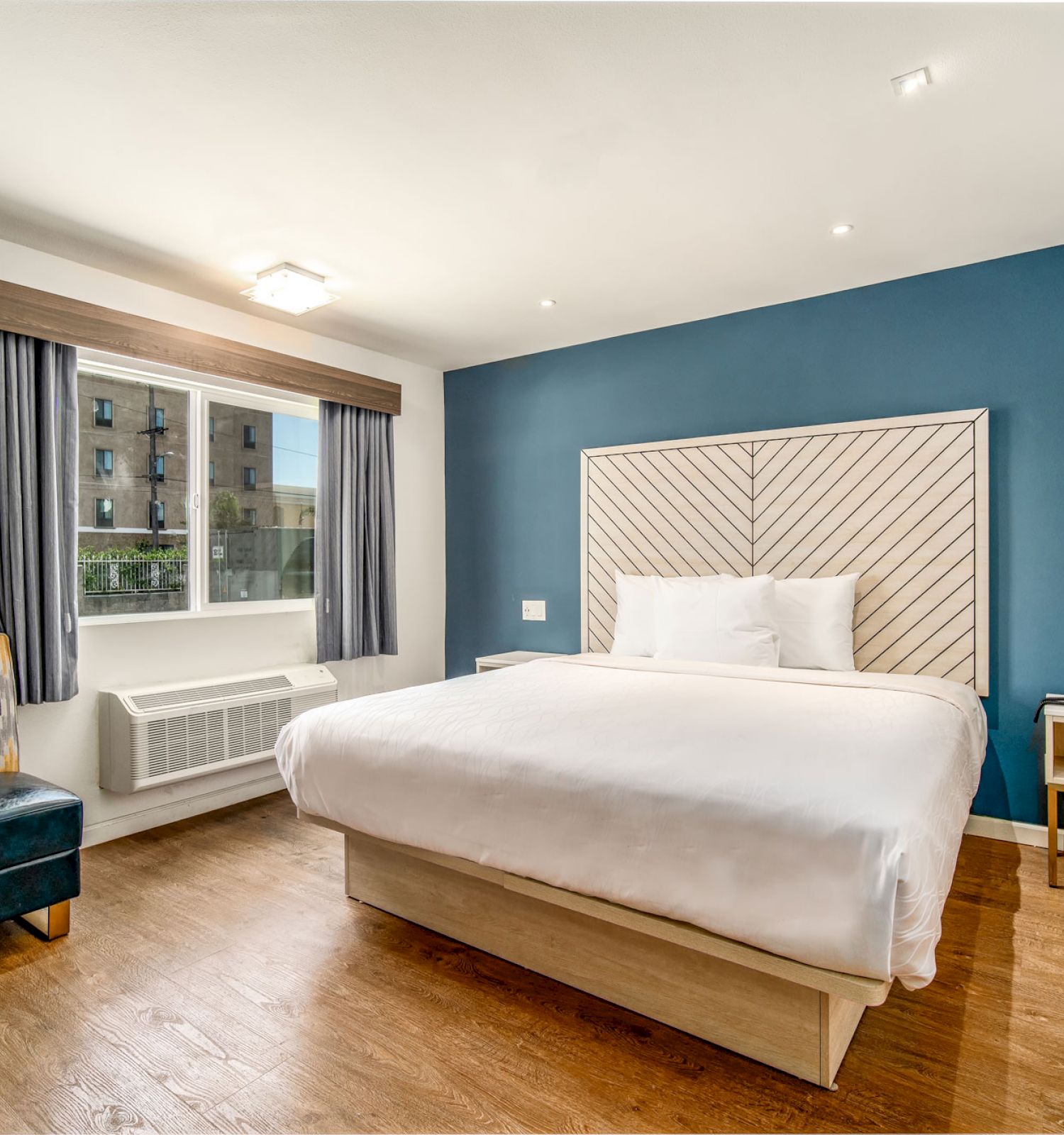 A cozy, modern hotel room with a large bed, stylish chair, window, and bedside table set against a blue accent wall and wood floor.