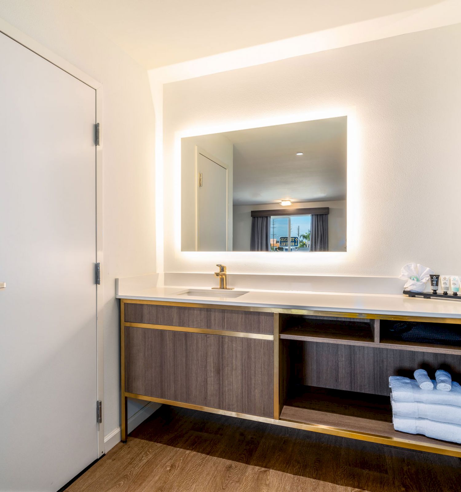 The image shows a bathroom vanity with a large, illuminated mirror, a sink, towels on a shelf, and toiletries on the counter.