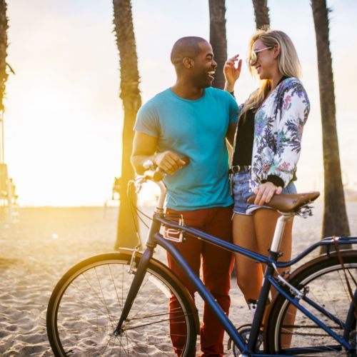 A couple enjoys a sunny beach with a bike, surrounded by palm trees. One person touches the other's face, both smiling in a warm moment.