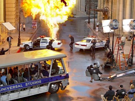 A movie set with an explosion, police cars, actors, and a film crew working while a Universal Studios tour bus passes by.