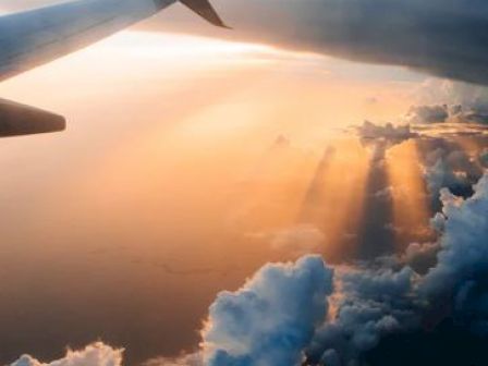 View from a plane window showing the wing, clouds below, and sunlight breaking through, creating a picturesque sky scene.